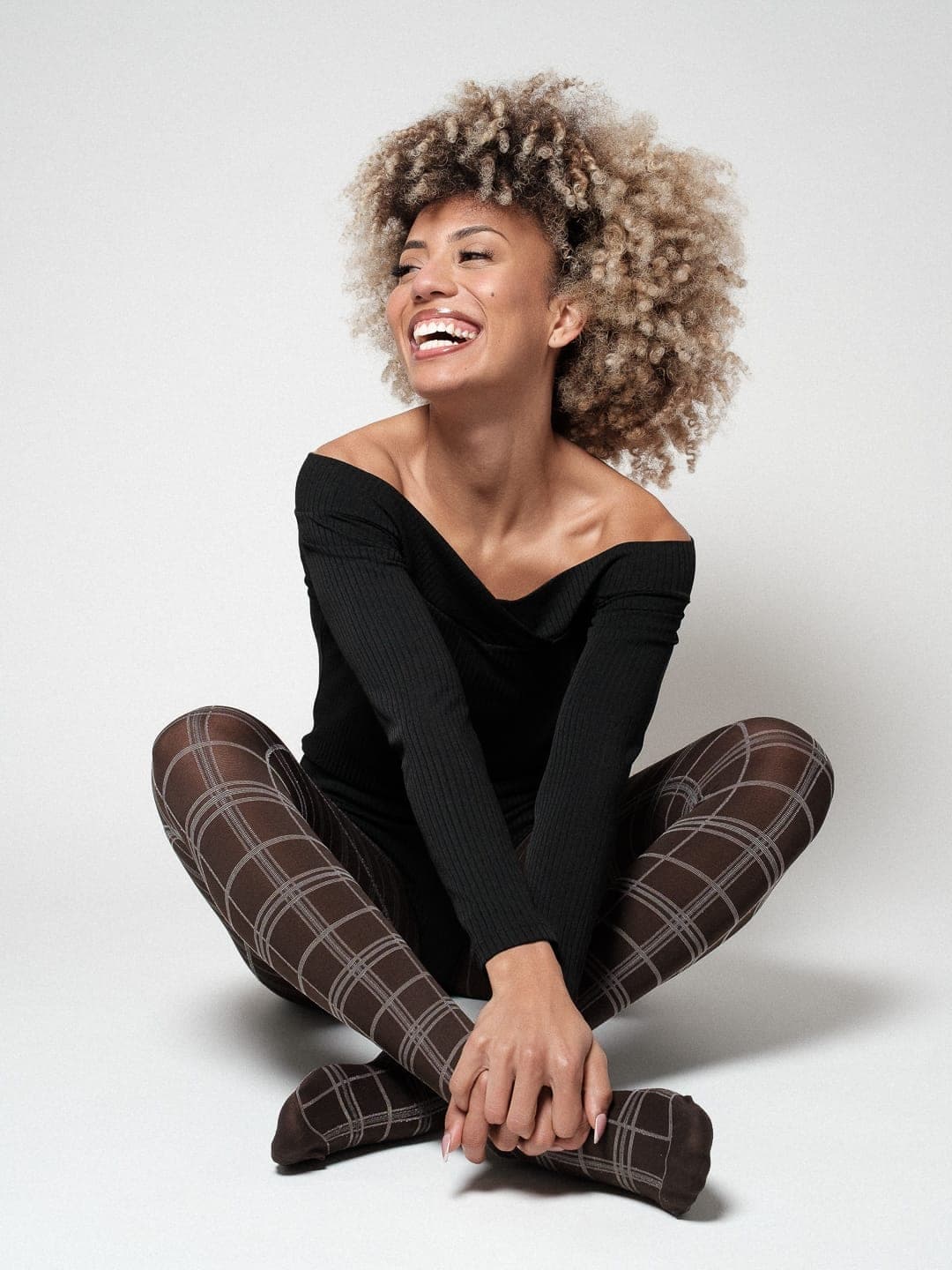 Tainá Ribeiro a rir, sentada de pernas cruzadas no chão do estúdio, em top preto de ombros descobertos e collants de xadrez